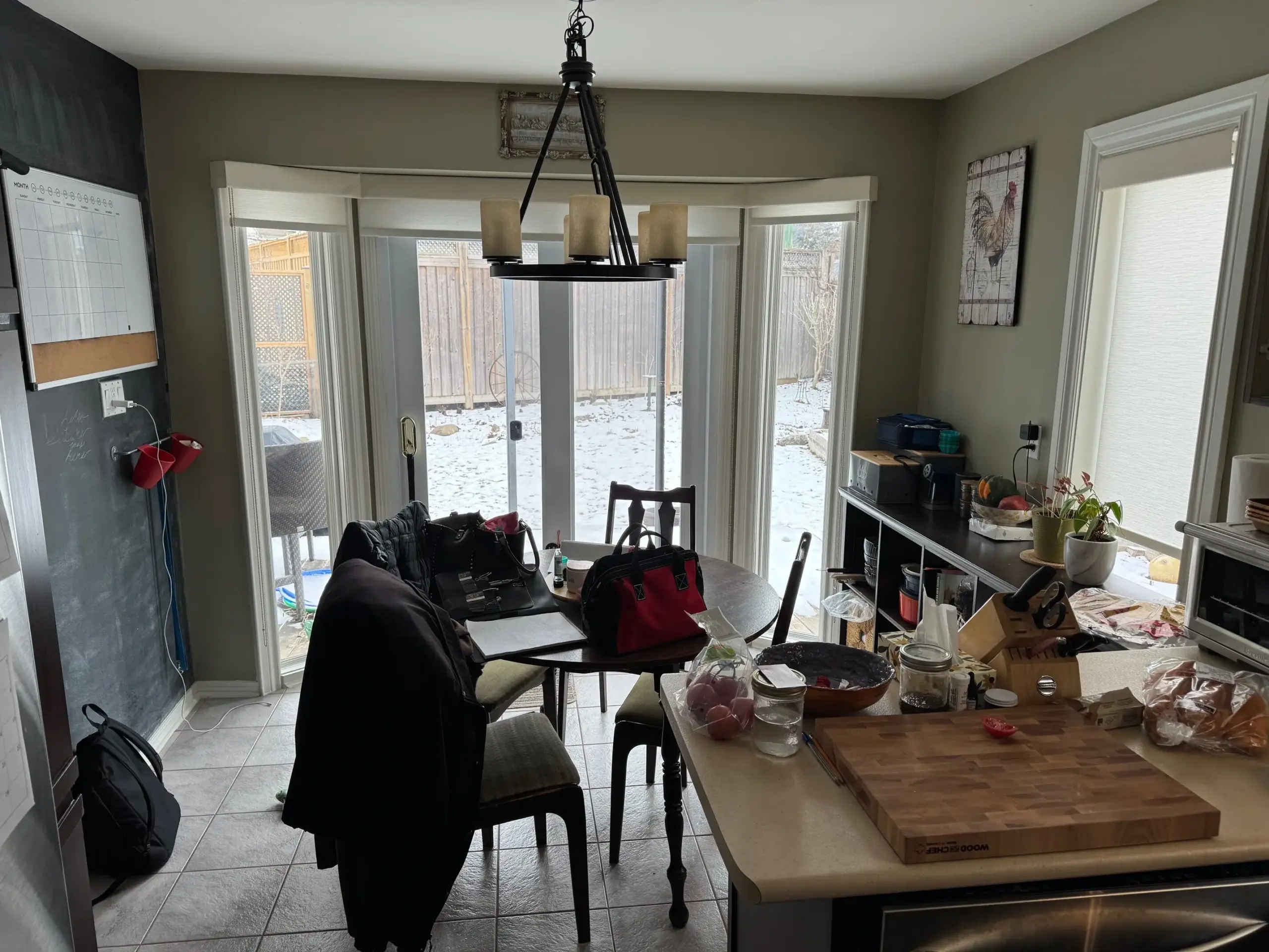 A cluttered kitchen and dining area with a round table, island, chandelier, and snowy backyard visible through large glass doors