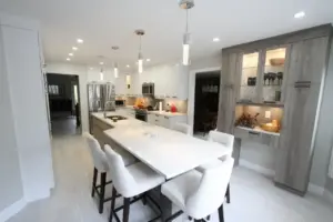 Modern white kitchen with a large island, four stools, pendant lights, stainless steel appliances, and gray built-in cabinets