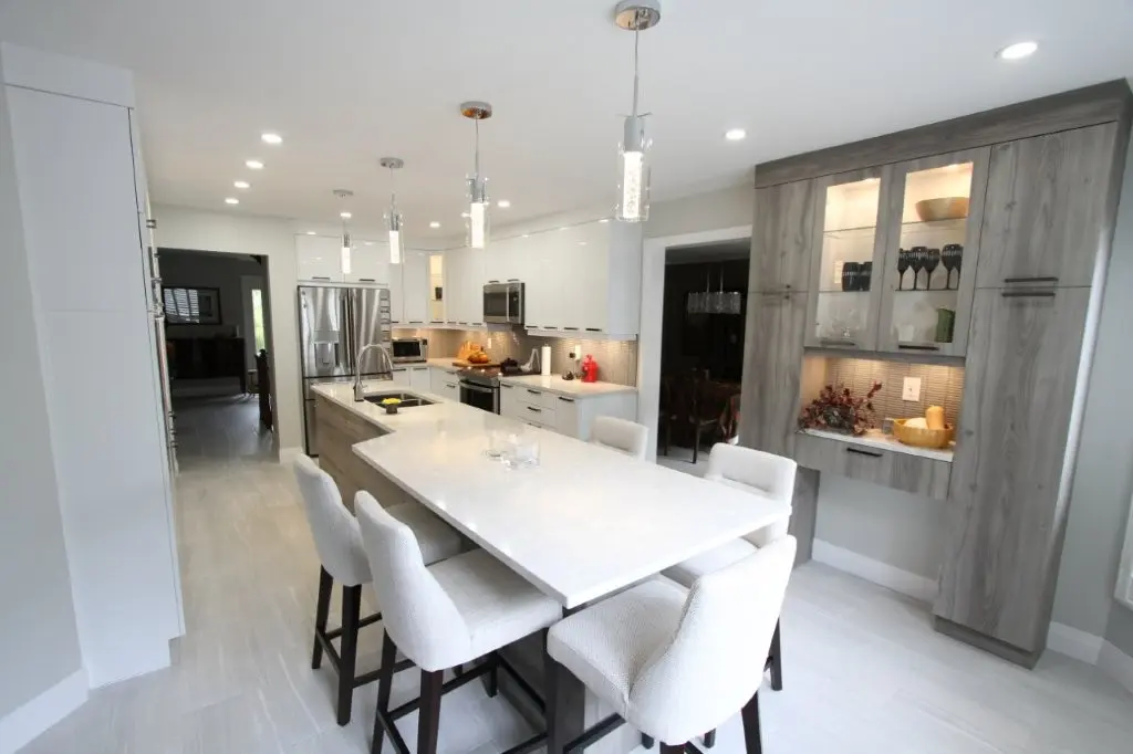 Modern white kitchen with a large island, four stools, pendant lights, stainless steel appliances, and gray built-in cabinets