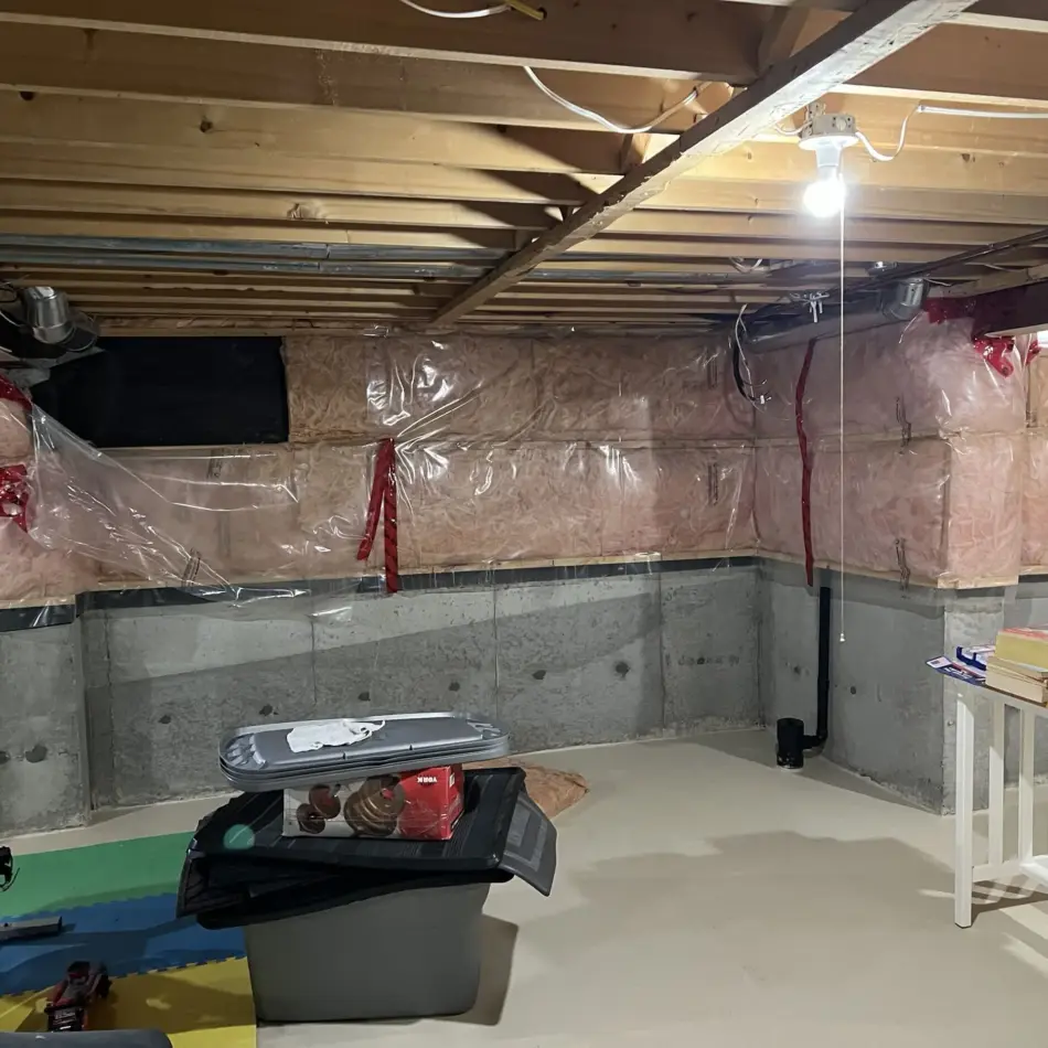 IMG_7682 Unfinished basement with exposed joists, insulated plastic-covered walls, and a single hanging bulb