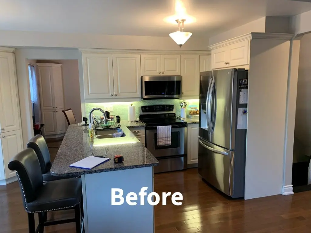 Kitchen before remodel with white cabinets, granite island, stainless appliances, and two black bar stools